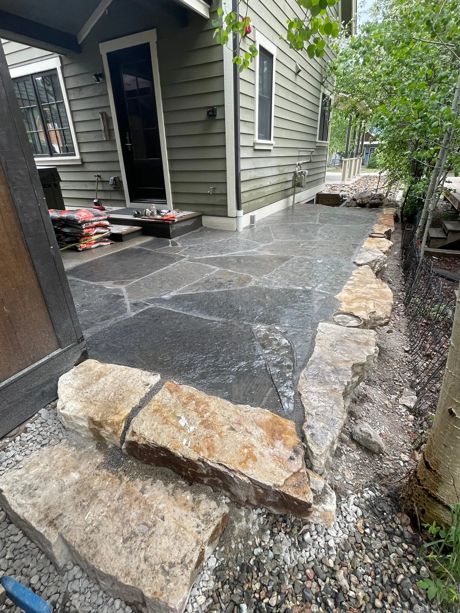 Flagstone patio installation in progress with boulder placement alongside a home